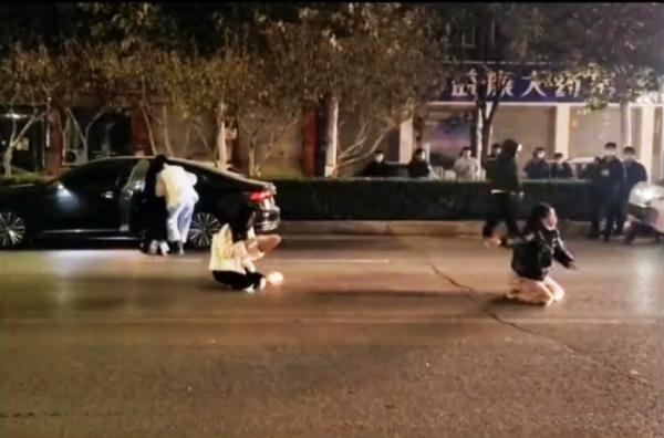 精神失常的女孩和母亲跪在马路上，女孩母亲向围观的人求助。